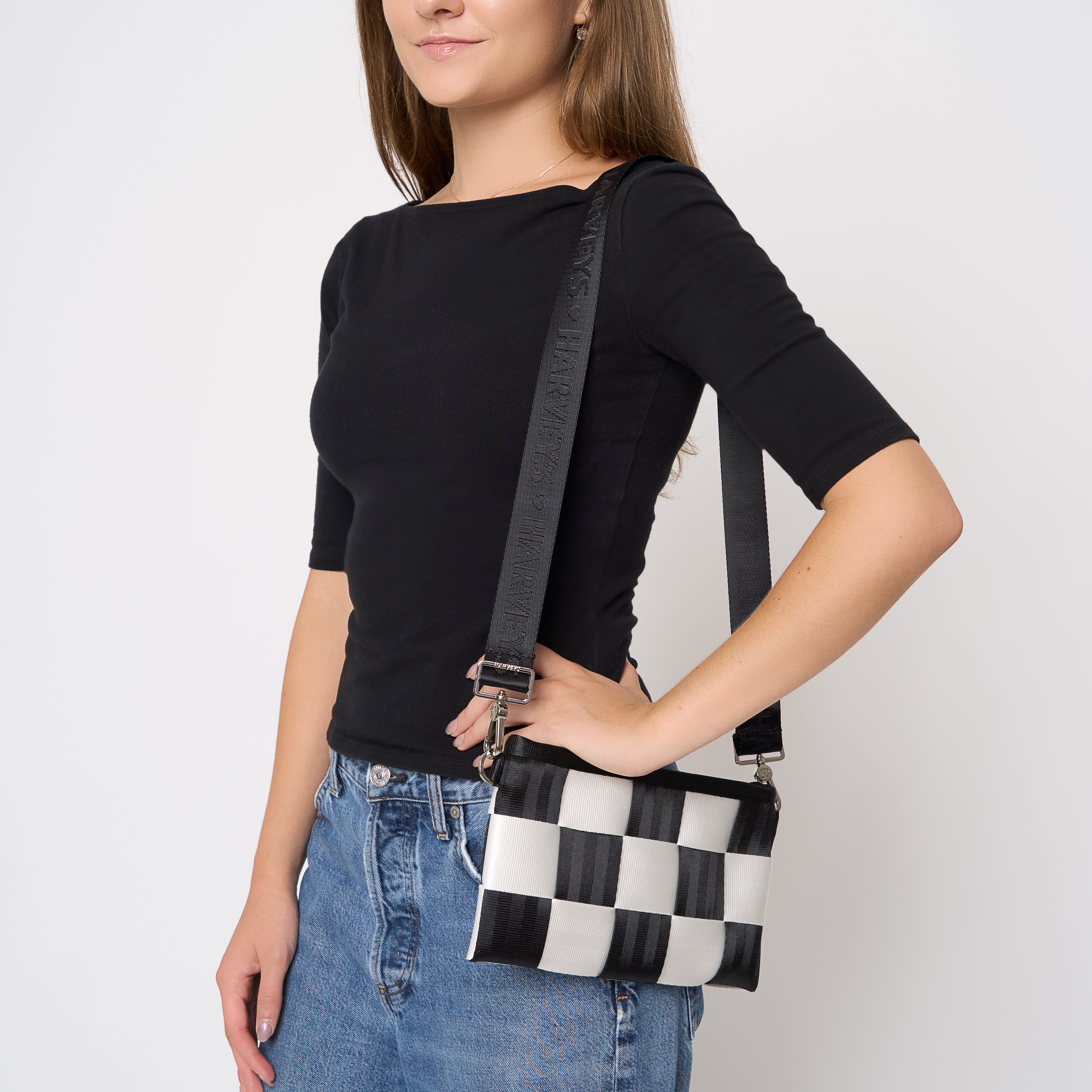 A woman wearing a black short-sleeve top and blue jeans carries on the shoulder a black and white checkered seatbelt crossbody bag with a black strap featuring white lettering, set against a plain white background.