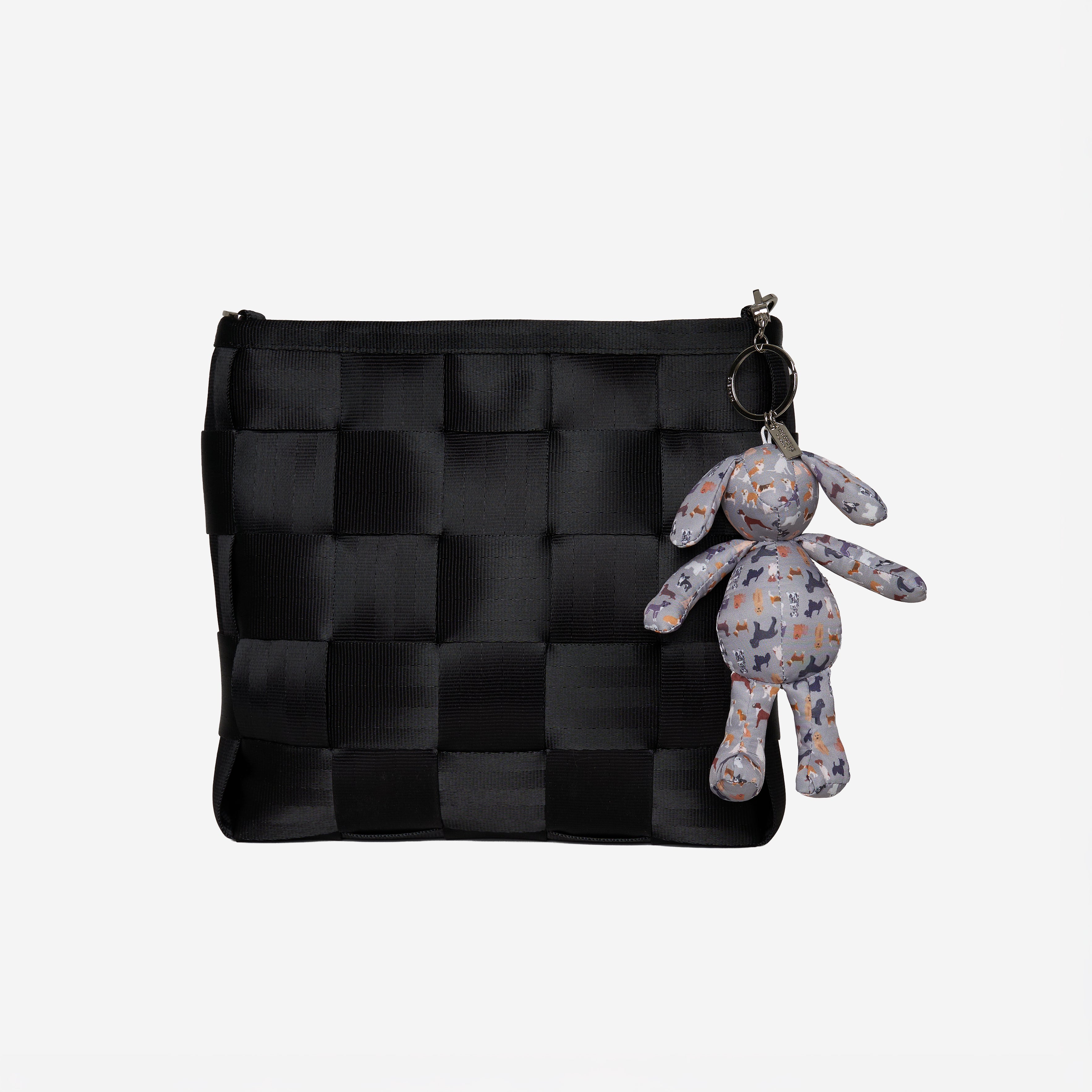A black woven mini messenger bag with a zipper closure, featuring a small gray plush dog keychain with a colorful pattern attached on a key ring, set against a plain light gray background.