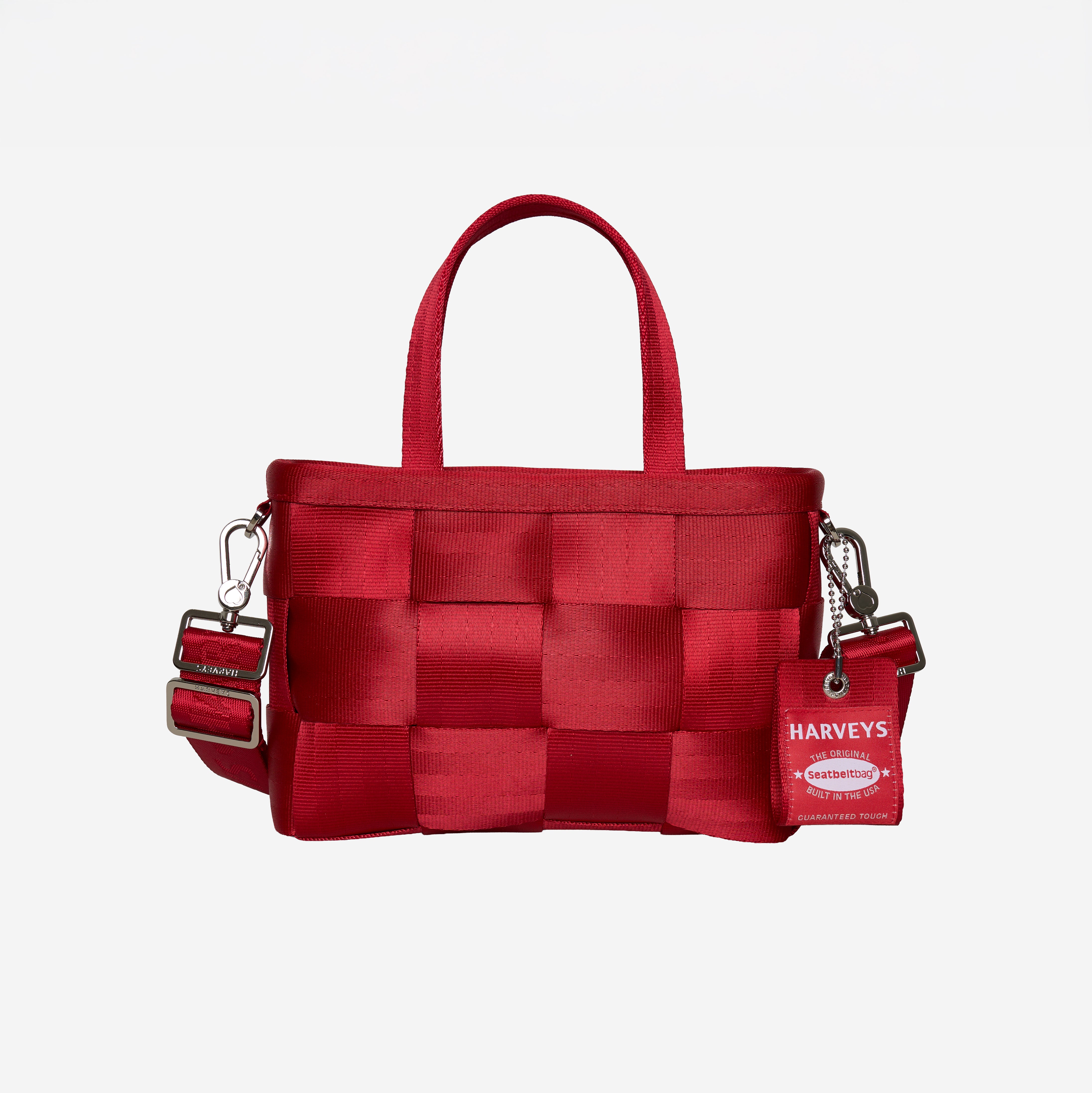 A red woven seatbelt handbag with top handles and an adjustable shoulder strap, featuring a hanging red tag with the brand name 'Harveys'.