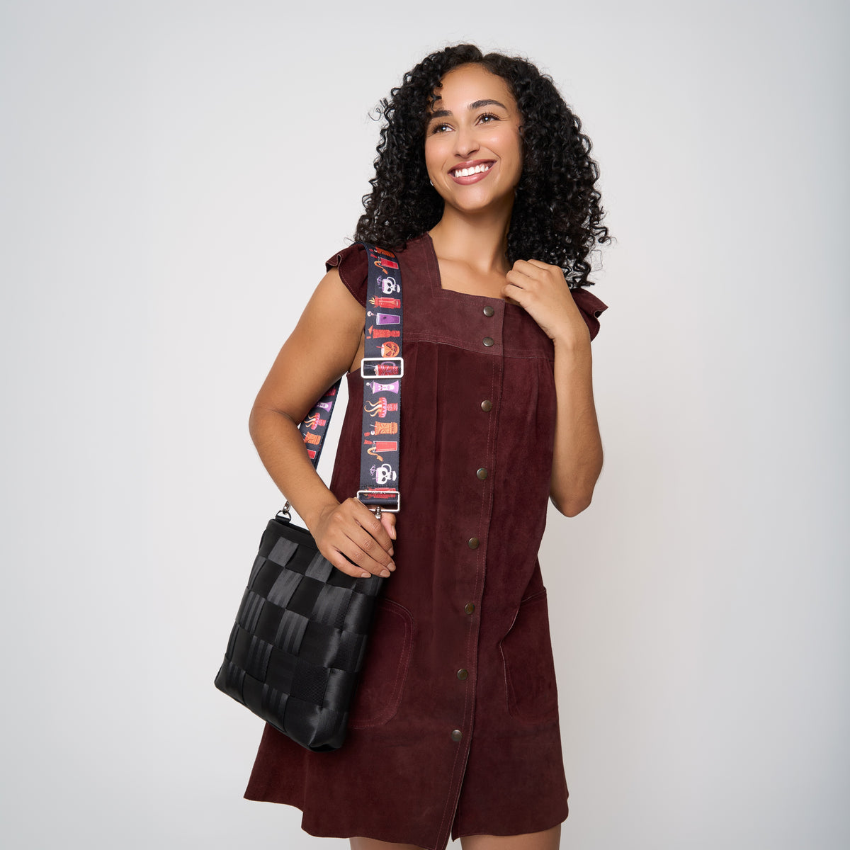 A woman with curly dark hair models a sleeveless burgundy dress with a button front and a black woven crossbody bag featuring Shag Spooky tiki cocktail patterned strap, set against a plain light gray background.