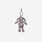 A small plush dog keychain with a multicolor abstract pattern on a light gray base, featuring a silver metal split ring and clasp at the top, shown against a plain light gray background.