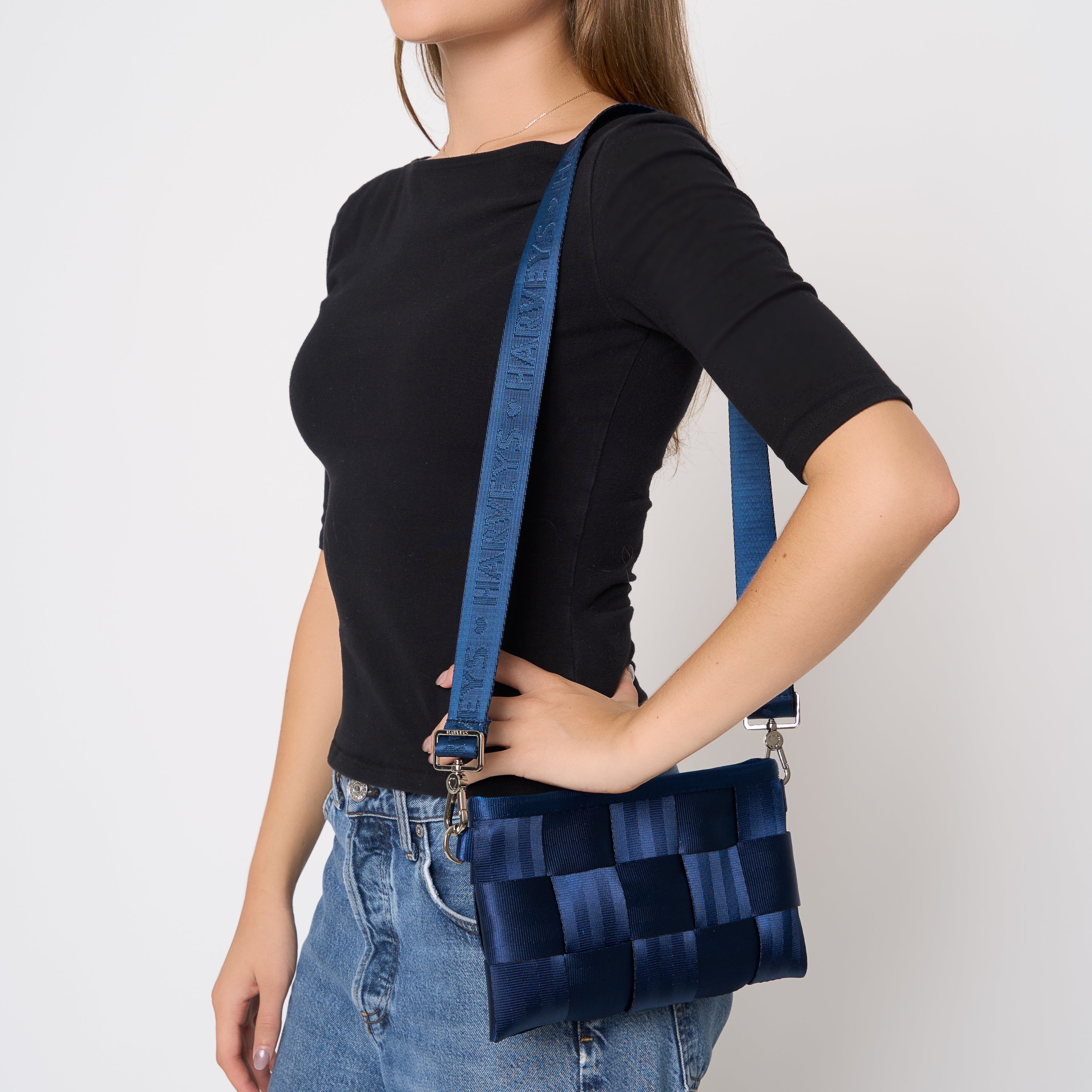 A woman wearing a black short-sleeve top and blue jeans carries a Indigo blue woven seatbelt crossbody Hip Pack bag with a matching woven strap. The bag has silver-tone hardware and is shown against a plain light gray background.