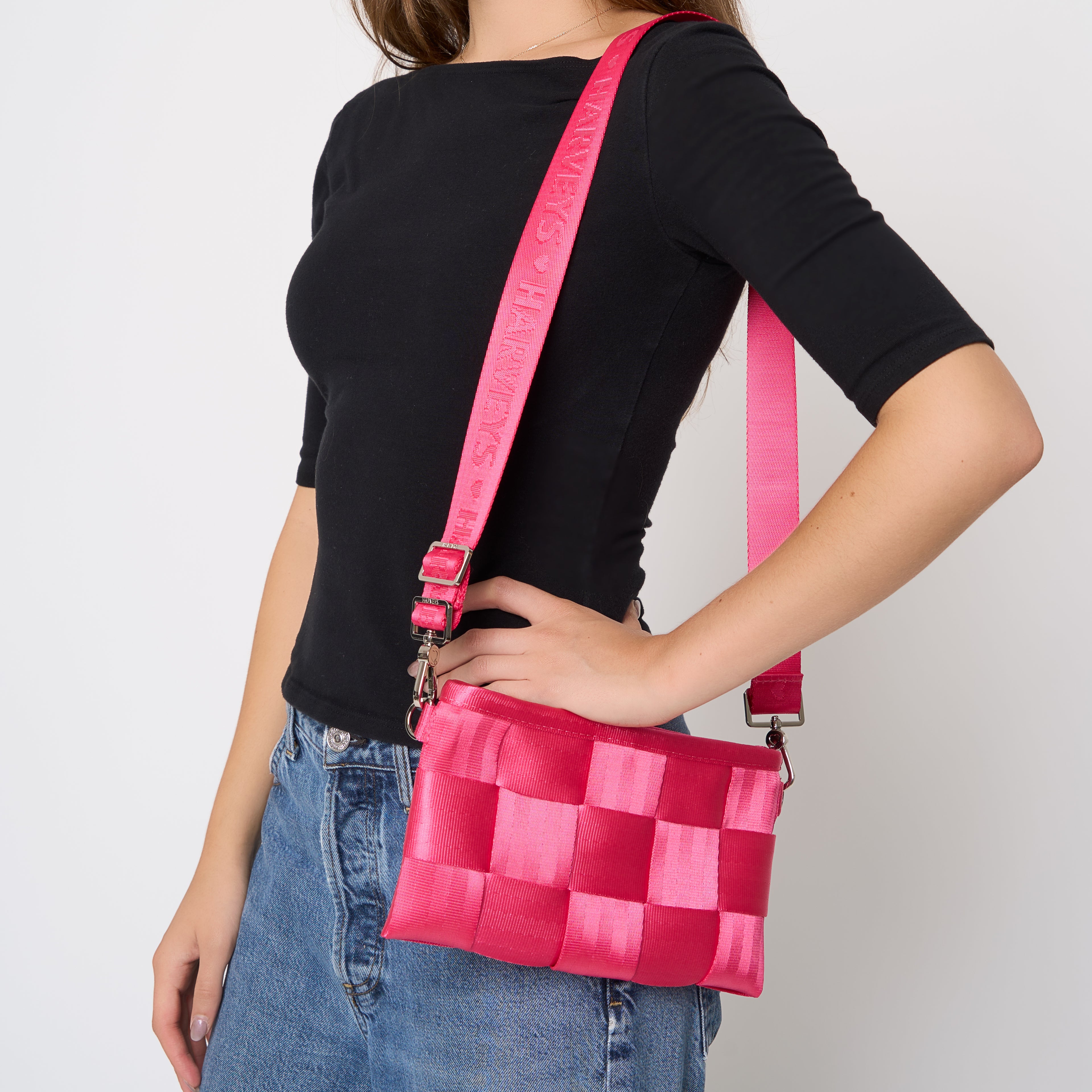 A woman wearing a black t-shirt and blue jeans carries a bright Melon pink woven crossbody bag with a matching pink strap set against a plain light gray background.