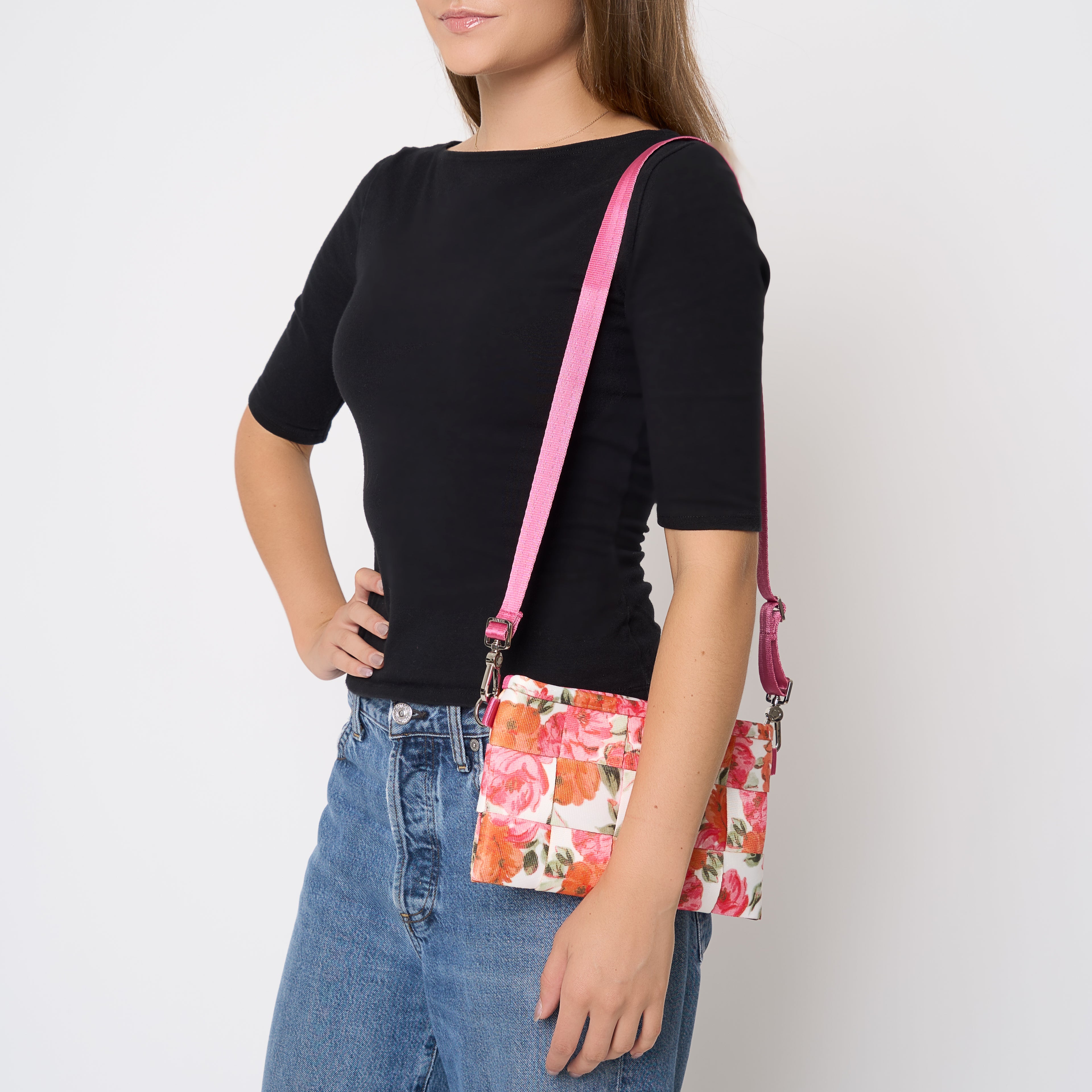 A woman wearing a black short-sleeve top and blue jeans models a Betty Rose floral seatbelt crossbody bag with a pink strap against a plain white background.