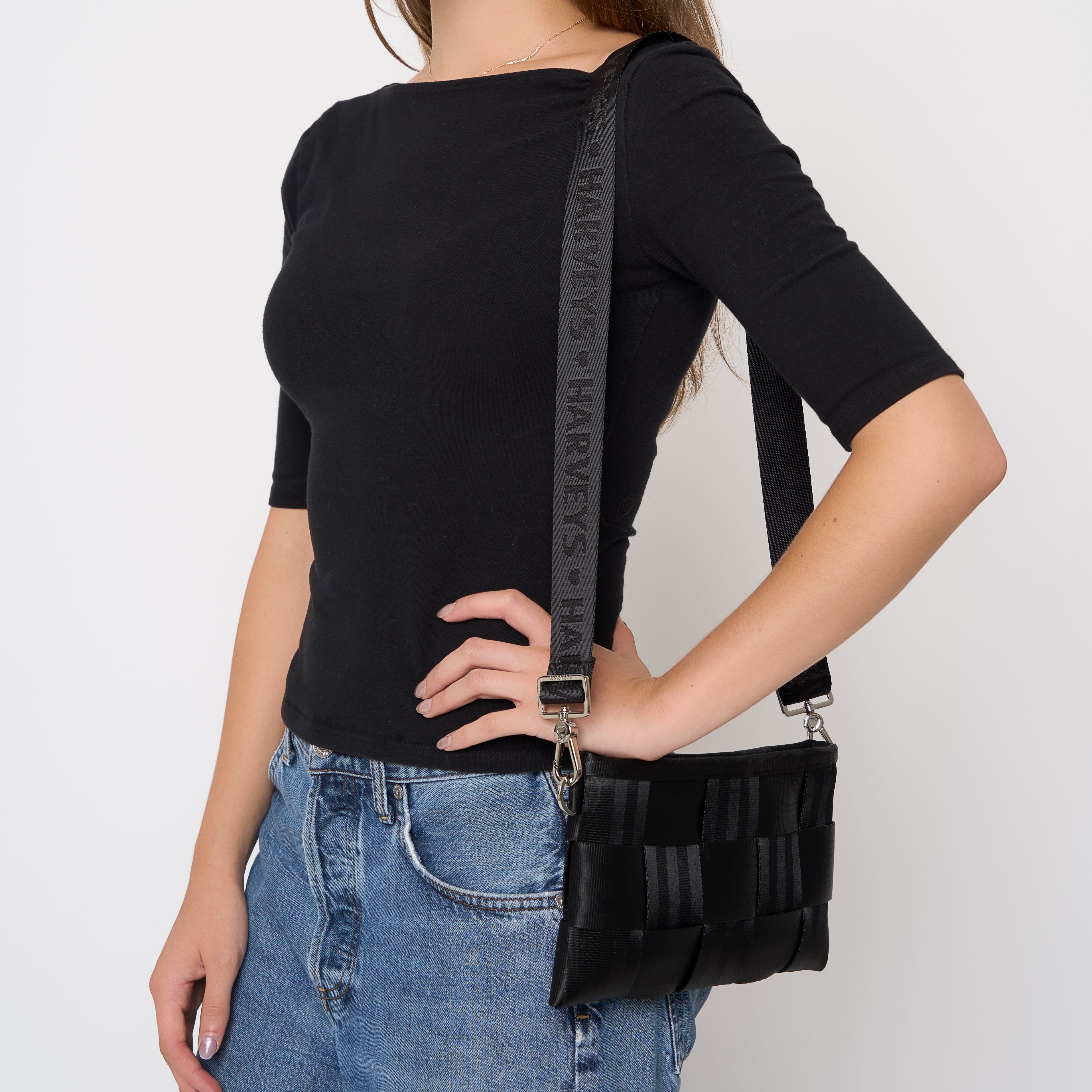 A woman wearing a black ribbed short-sleeve top and blue jeans carries a black woven seatbelt crossbody bag with a black Harveys branded strap against a light gray studio background.