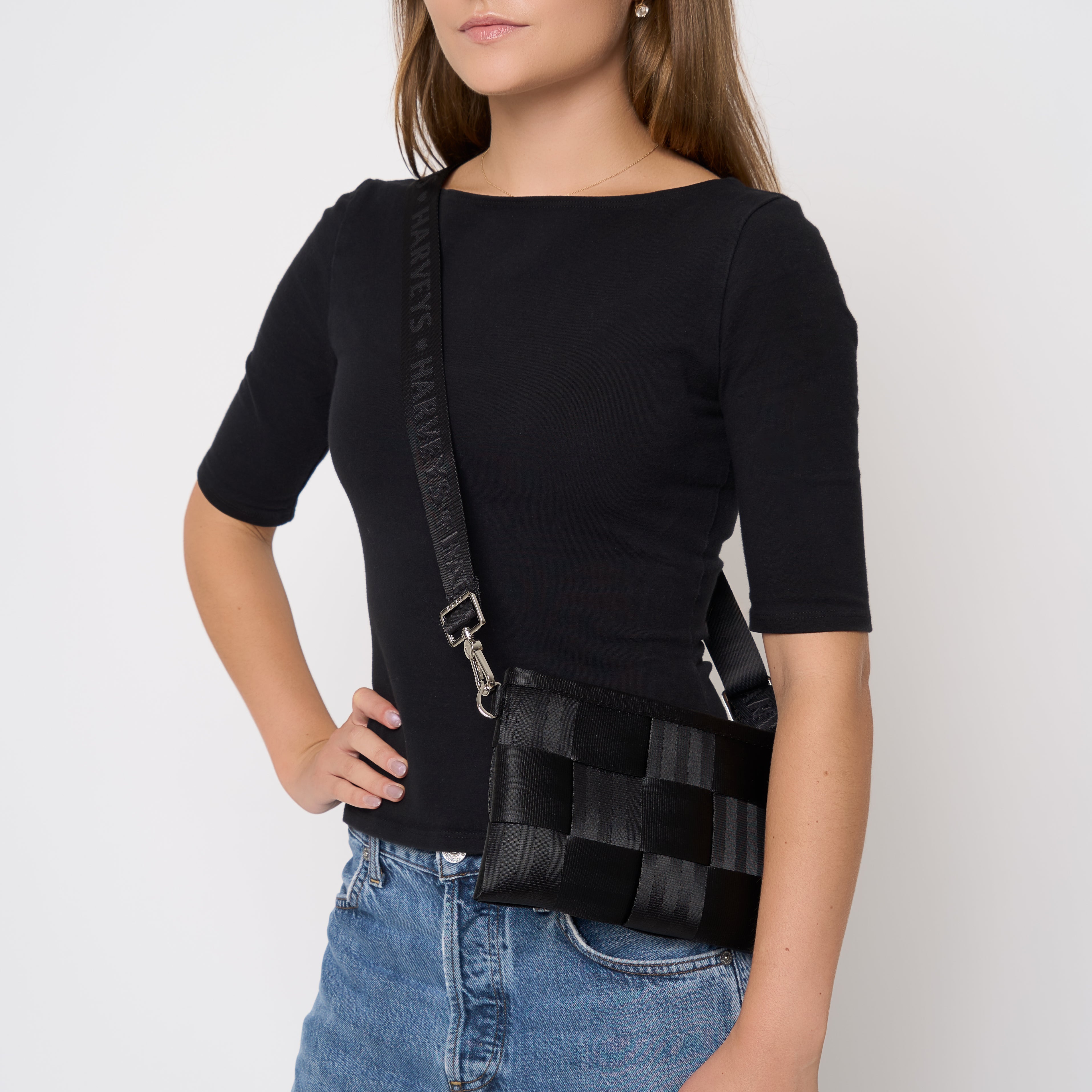 A woman wearing a black short-sleeve top and blue jeans models a black woven seatbelt crossbody bag with a wide strap and silver hardware against a plain light gray background.