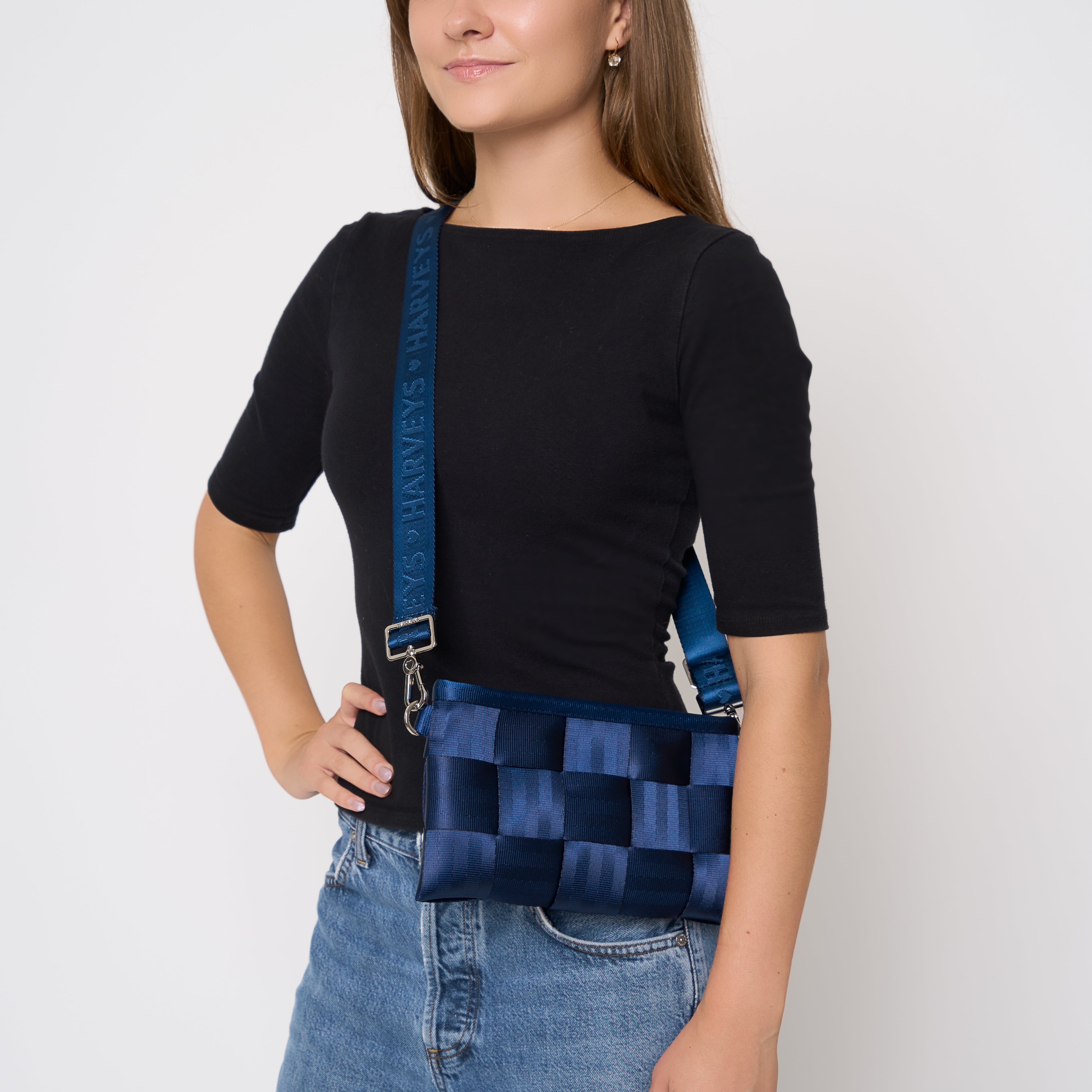 A woman wearing a black short-sleeve top and blue jeans models an Indigo blue woven seatbelt crossbody Hip Pack bag with a matching strap.