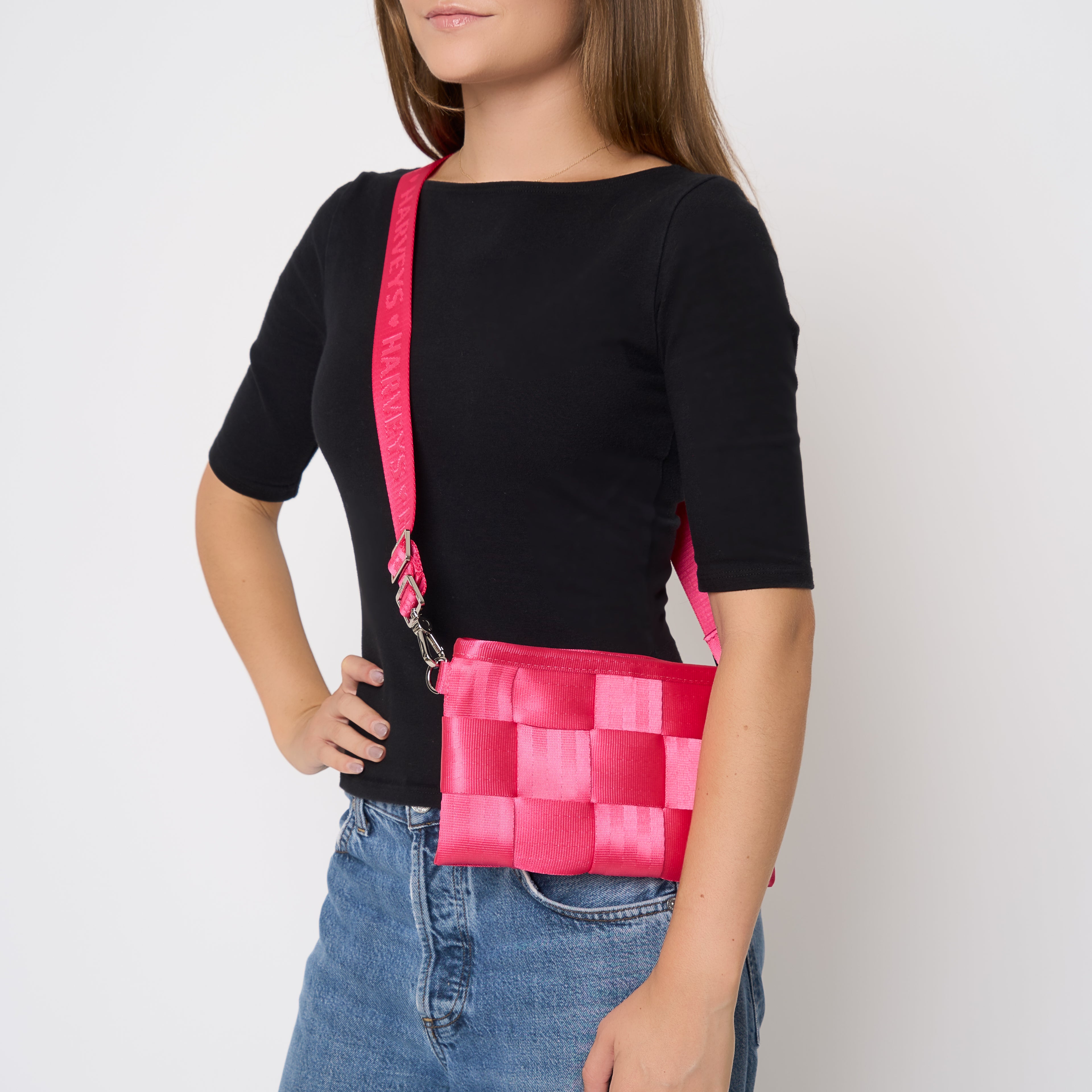A woman wearing a black short-sleeve top and blue jeans models a Melon pink woven crossbody bag with a matching pink strap against a plain light gray background.