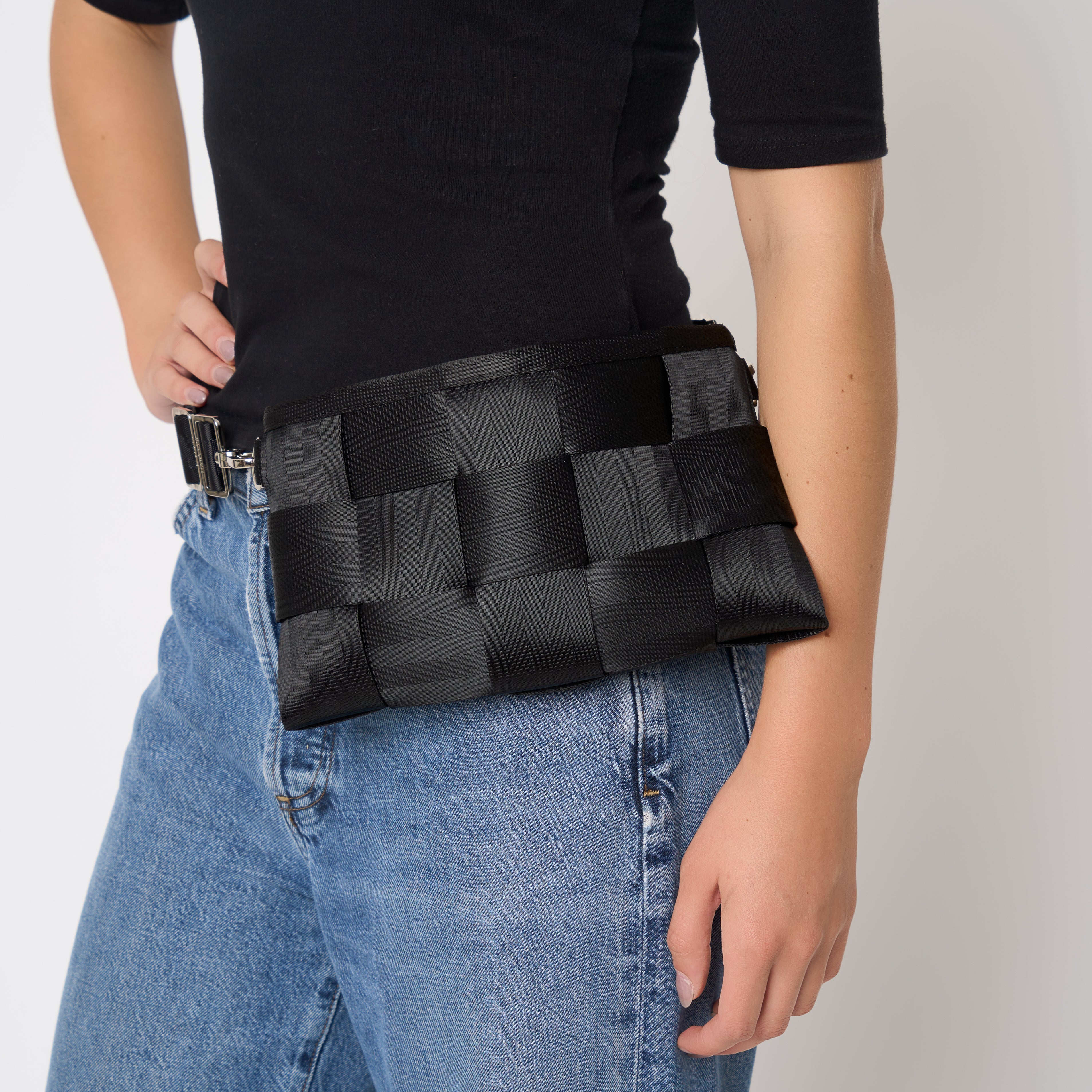A person wearing a black woven seatbelt bag with a black belt worn around the waist, styled with a black t-shirt and blue jeans against a light gray background.