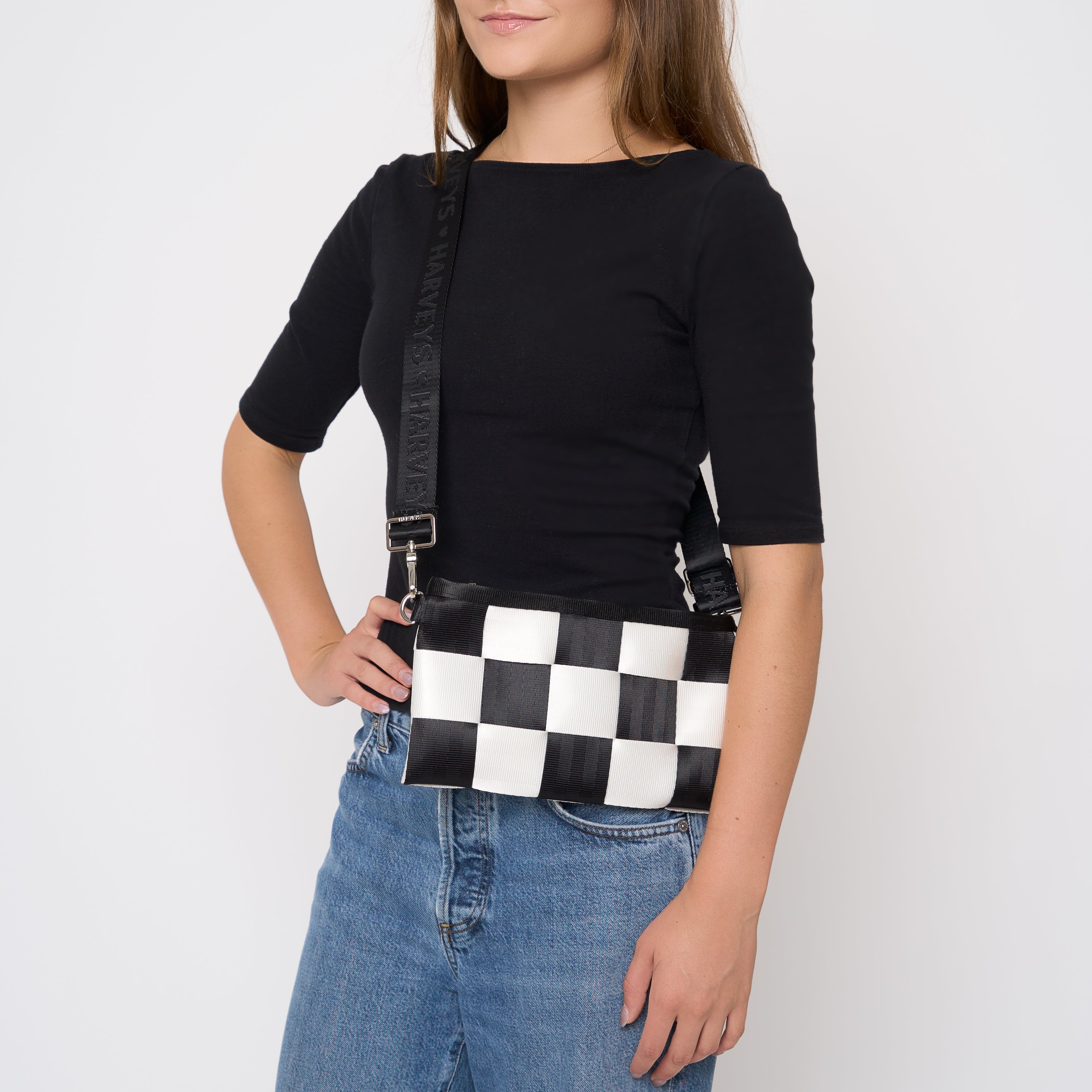 A woman wearing a black t-shirt and blue jeans models a black and white checkered seatbelt crossbody bag with a black strap.