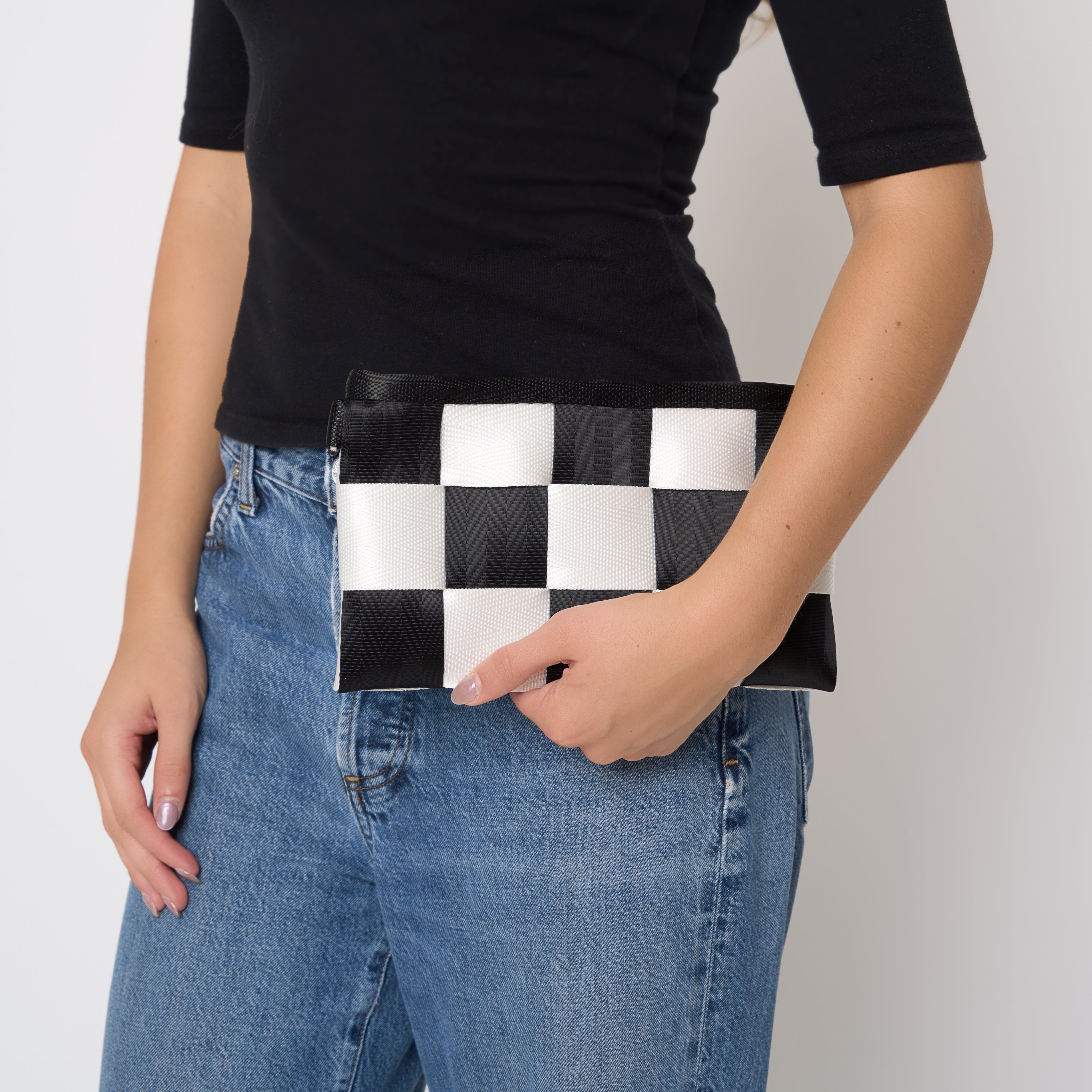 A person wearing a black t-shirt and blue jeans holds a seatbelt black and white checkered Hip Pack bag against their hip, shown as a clutch purse against a plain light gray background.