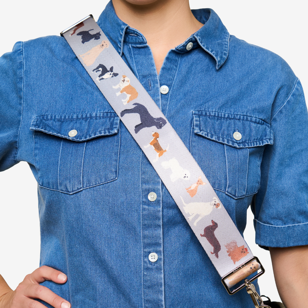A close-up of a person wearing a blue denim shirt and carrying a shoulder bag with a wide, light gray strap printed with various dog illustrations in black, white, orange, and brown.