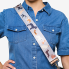 A close-up of a person wearing a blue denim shirt and carrying a shoulder bag with a wide, light gray strap printed with various dog illustrations in black, white, orange, and brown.