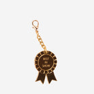 A gold-tone metal keychain or bag charm shaped like a 'Best in Show' award ribbon badge, featuring a lobster clasp and the words 'BEST IN SHOW' printed in gold on a dark brown center, set against a plain light gray background.