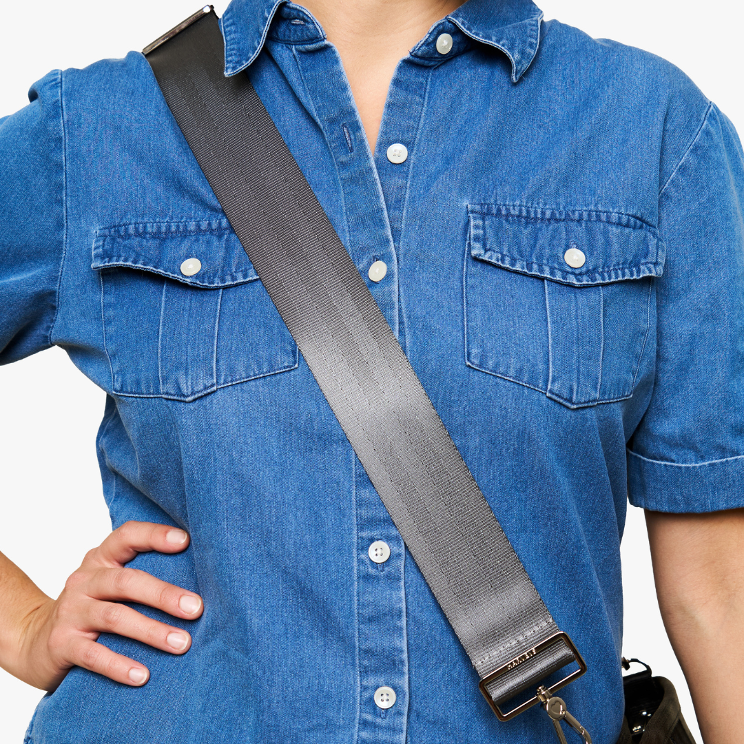 A close-up of a person wearing a blue denim shirt, showing a 5 Panel Click n Carry Storm shoulder strap with a metal buckle crossing the chest. The person's hand is resting on their hip, and a small black bag is partially visible at the bottom right.